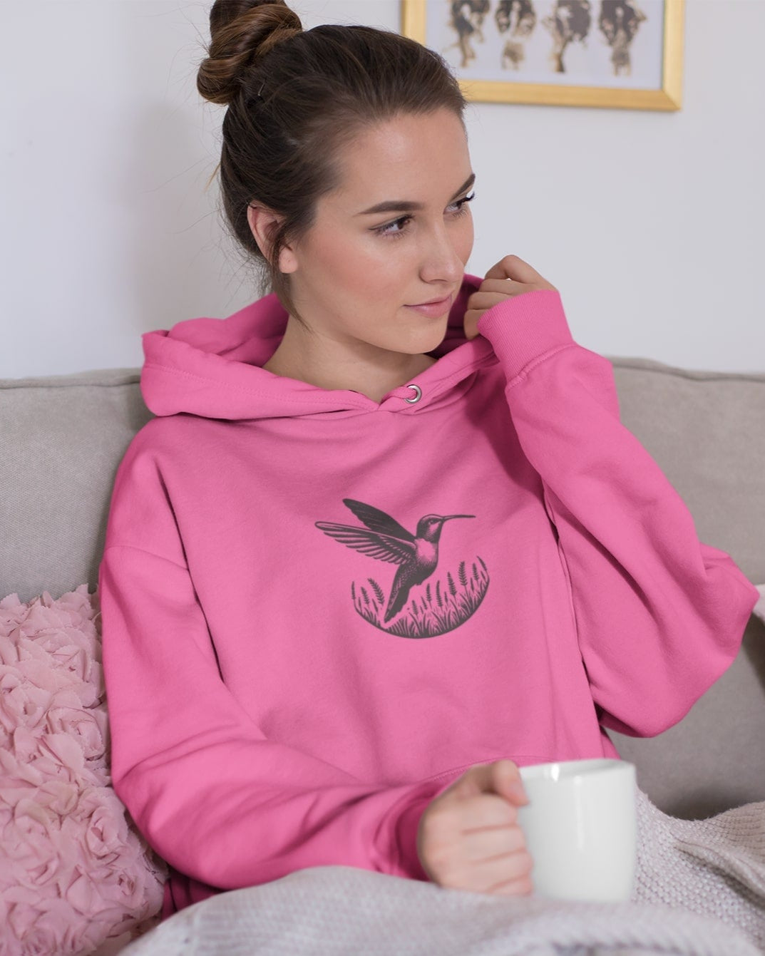 Woman wearing a pink hoodie with a bird design, sitting on a couch holding a mug.